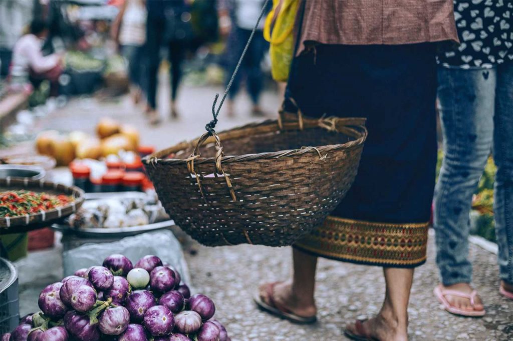 picture of the bottom half of a lady as she carries and empty basket. Some vegetables are in a heap next to her, to be sold.